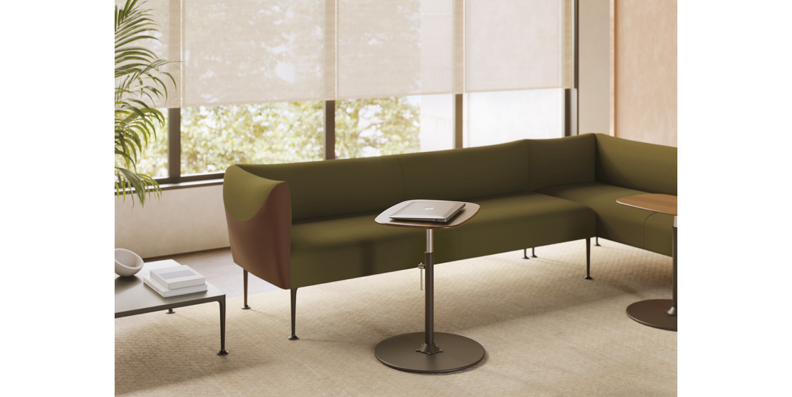 Lounge area with an olive green Missiva sofa in a corner layout and a Vetona height-adjustable side table holding a laptop